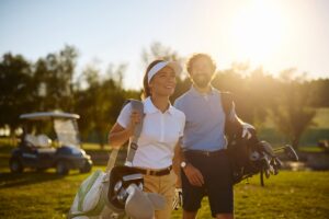 A man and a woman carrying golf bags walk together across a sunlit golf course at sunset.